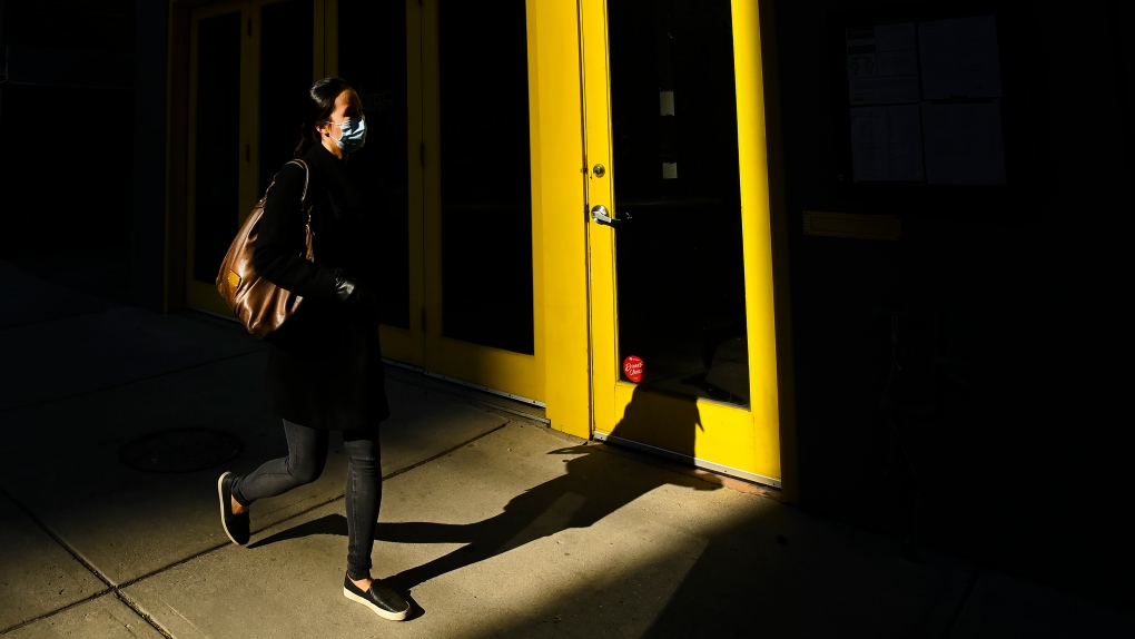 A woman wearing a protective mask walks through a shaft of light during the COVID-19 pandemic in downtown Toronto on Thursday, November 12, 2020 (The Canadian Press/Nathan Denette).
